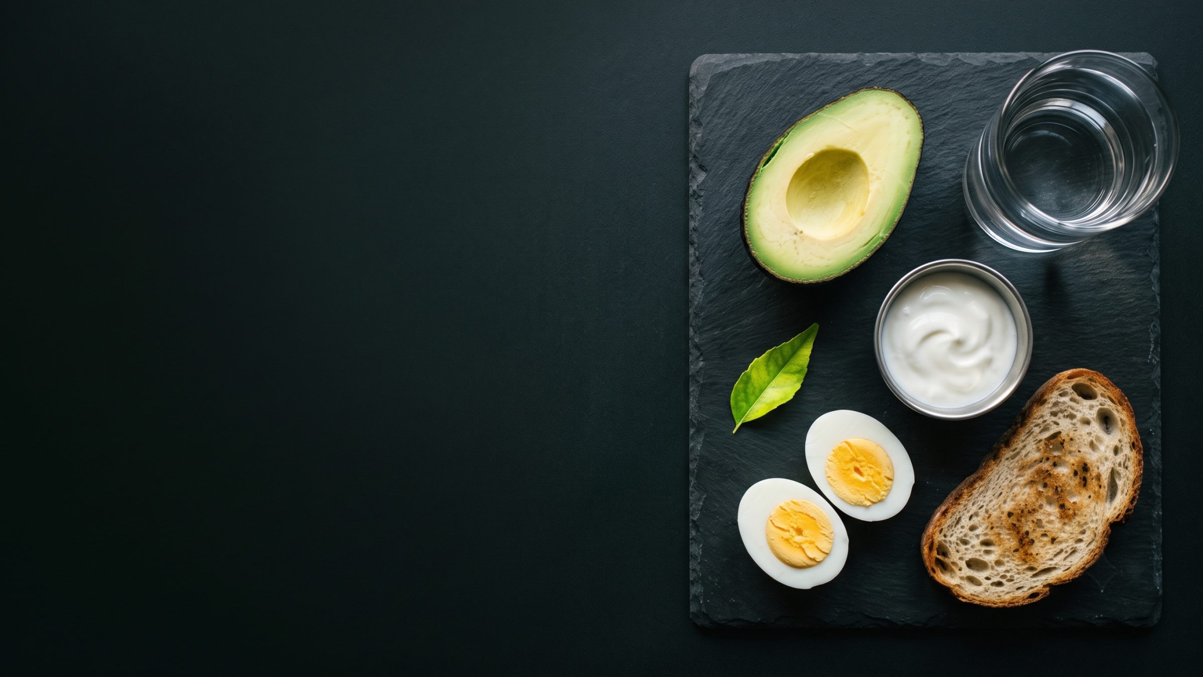 Half an avocado with boiled eggs, yoghurt and toast on a dark slate board